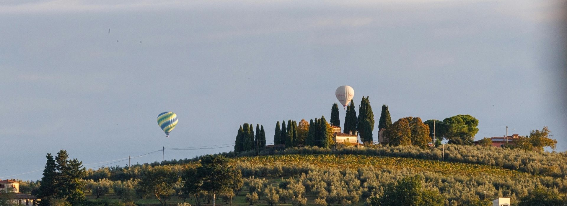 Due mongolfiere in volo all'alba sopra una collina toscana con vigneti, ulivi, cipressi e un casale rurale immerso nella campagna del Chianti