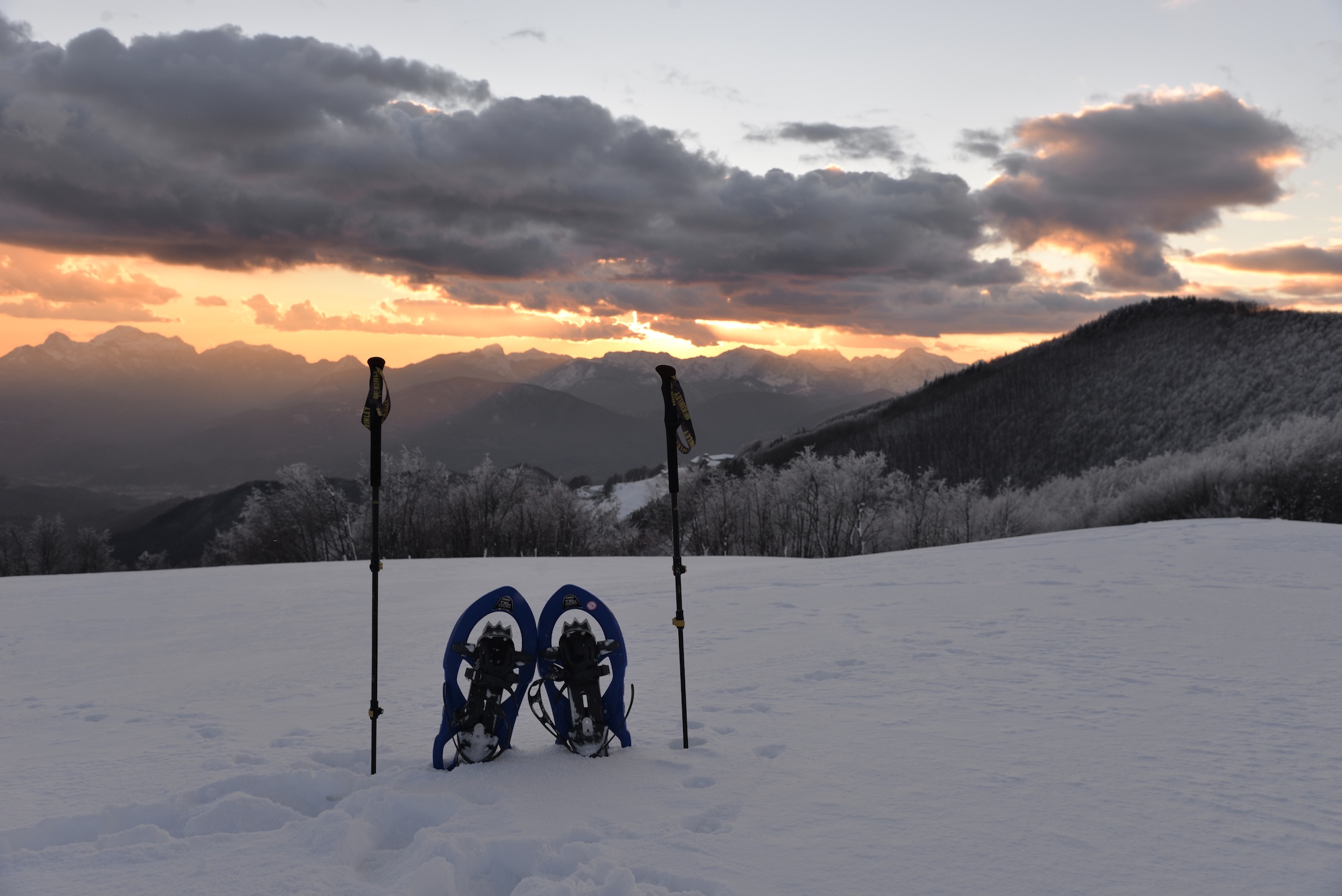 Raquettes et bâtons bleus plantés dans la neige sur une pente enneigée ; en arrière-plan, les montagnes de la Garfagnana au coucher du soleil