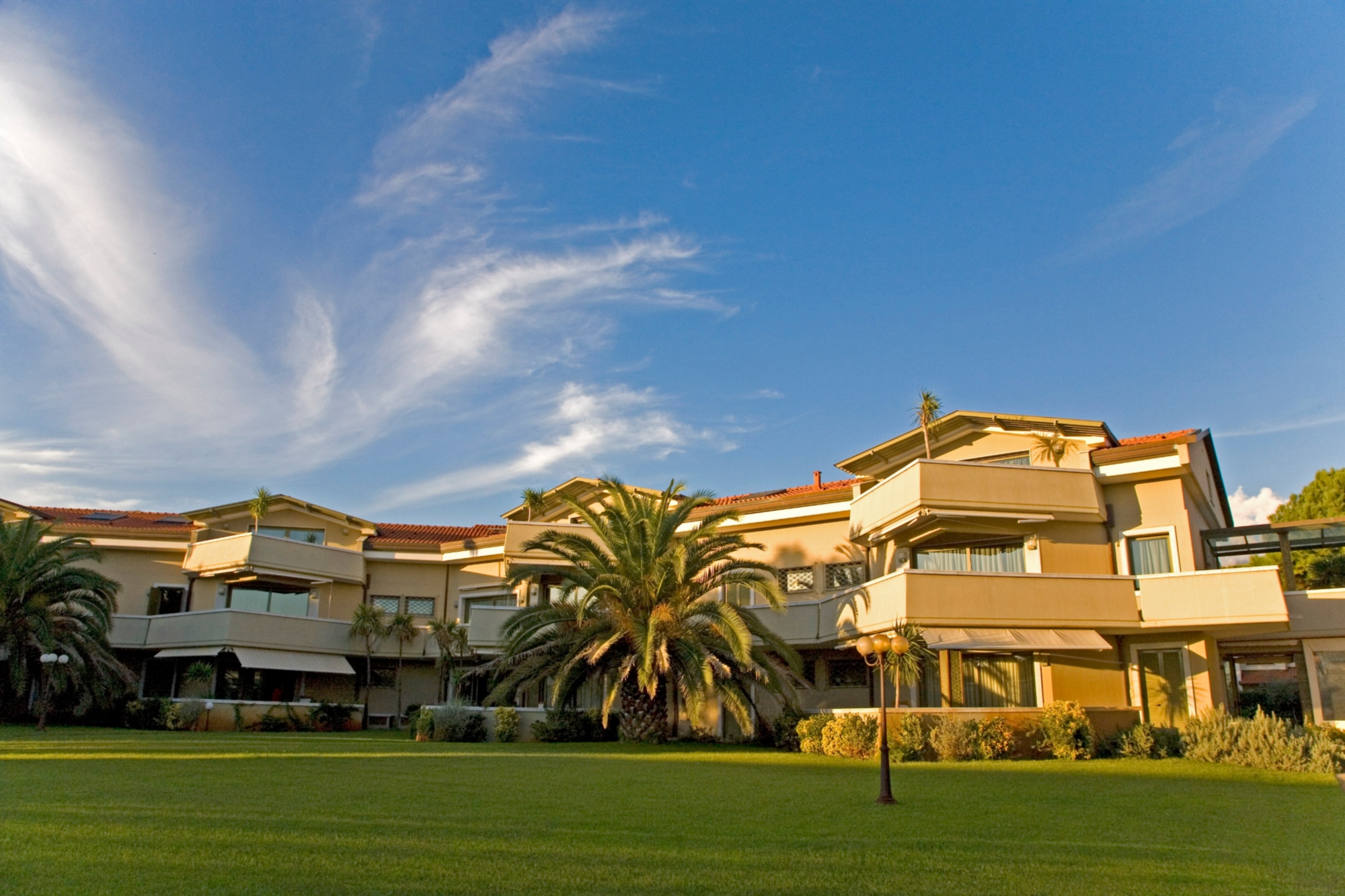 Facciata esterna delle Terme della Versilia con giardino curato, ampio prato verde, palme e balconi delle camere, cielo azzurro al tramonto