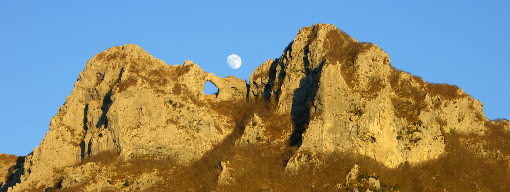 Vette rocciose del Monte Forato nelle Alpi Apuane illuminate dalla luce calda del tramonto, con il caratteristico arco naturale in pietra visibile tra le due cime e la luna che sorge sullo sfondo di un cielo azzurro limpido