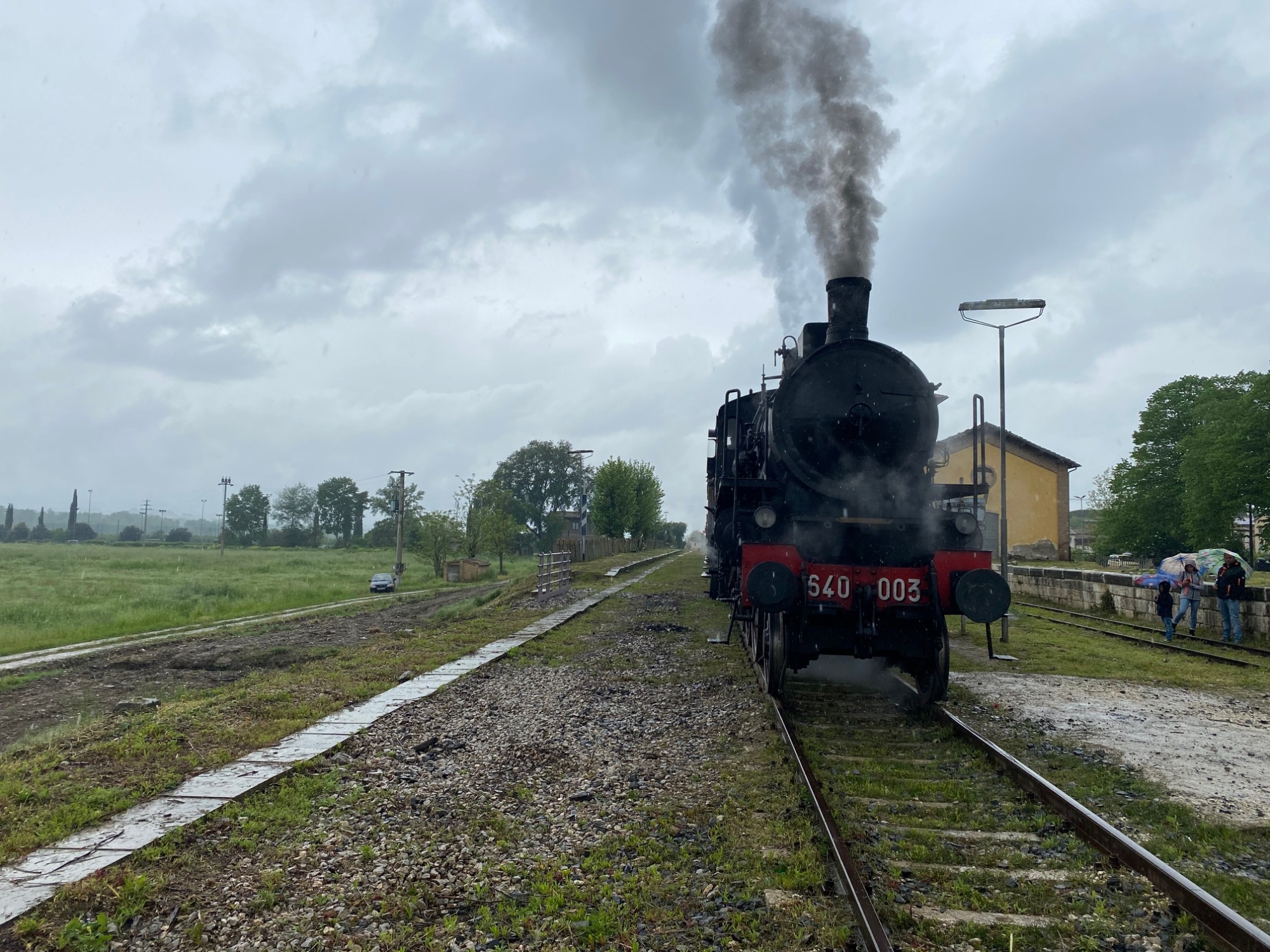 Locomotiva a vapore 640-003 del Treno Natura ferma sui binari di una piccola stazione toscana, con il fumo nero che sale dalla ciminiera e il cielo nuvoloso sullo sfondo, durante l'escursione storica da Siena ad Abbadia San Salvatore