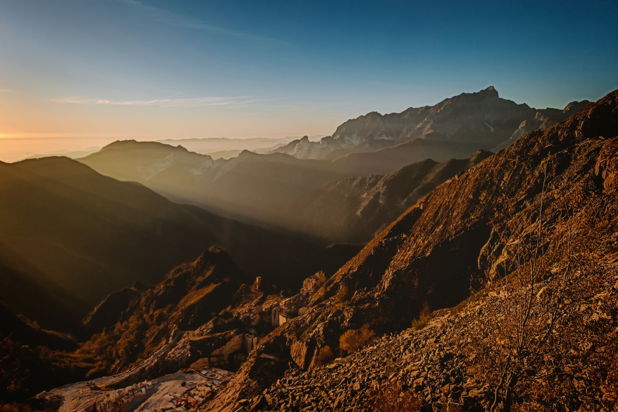 Panorama montano delle Alpi Apuane al tramonto, con i raggi del sole che filtrano tra i versanti rocciosi illuminando il paesaggio, una cava di marmo nella valle sottostante e vette che si profilano all'orizzonte sotto un cielo limpido