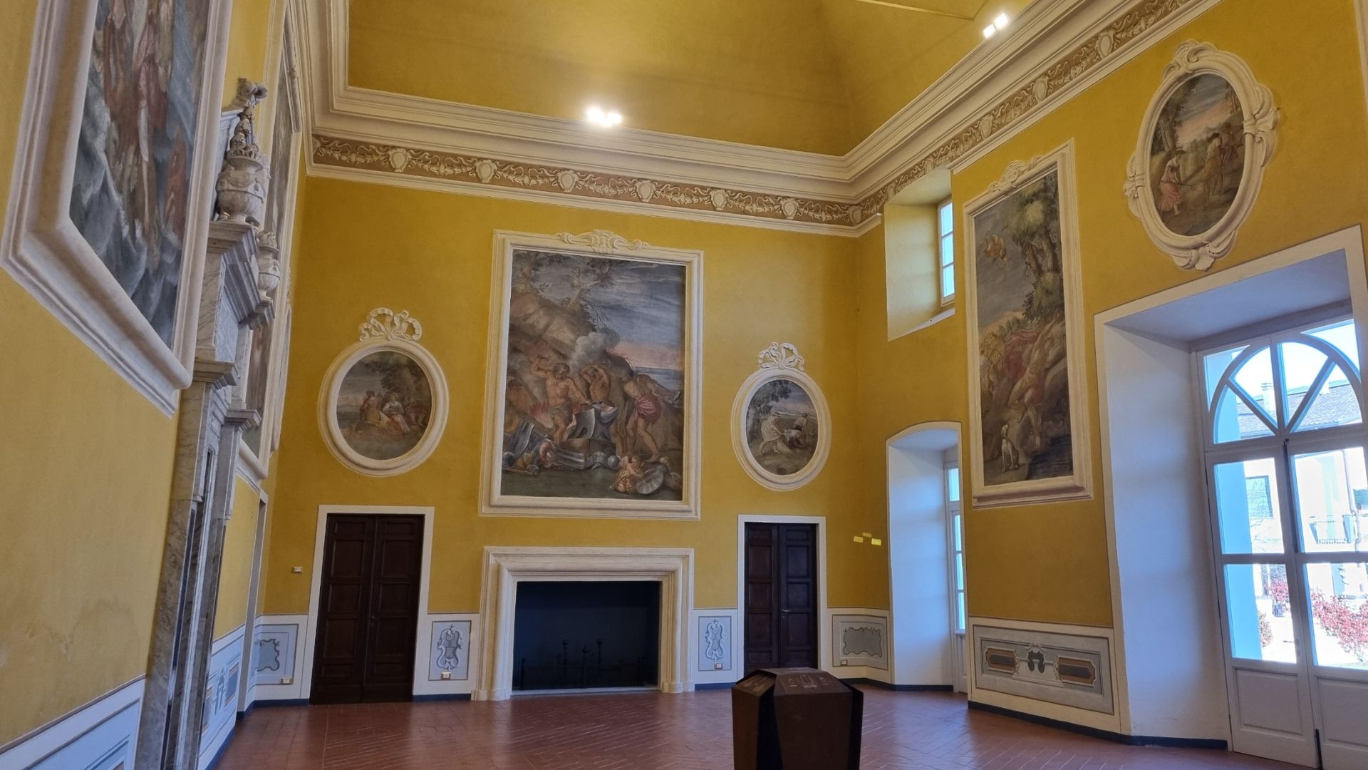 Salone affrescato del Castello di Pallerone con pareti gialle, grandi dipinti storici, camino centrale e finestre ad arco.