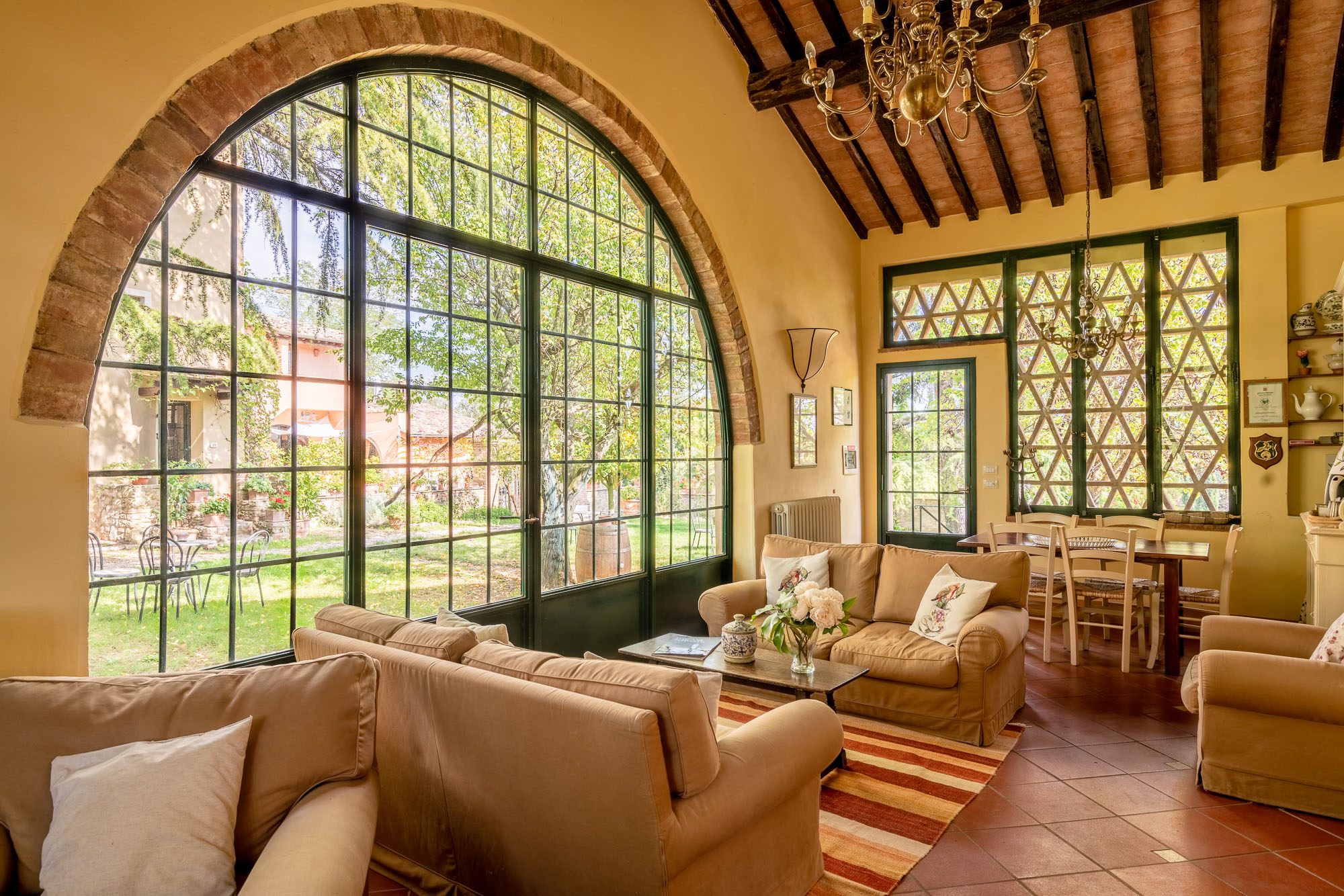 Ampio salone della Torraccia di Chiusi con divani beige, grande vetrata ad arco con vista sul giardino, soffitto con travi in legno a vista, lampadario in ottone e pavimento in cotto toscano