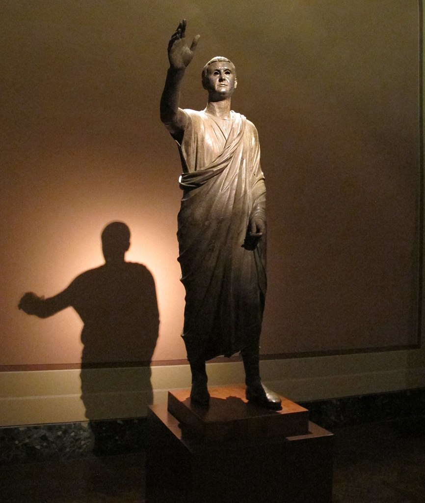 Statue romaine en bronze connue sous le nom de L’Arringatore, représentant un orateur debout, le bras droit levé, symbole d’éloquence et d’autorité, conservée au Musée archéologique national de Florence.