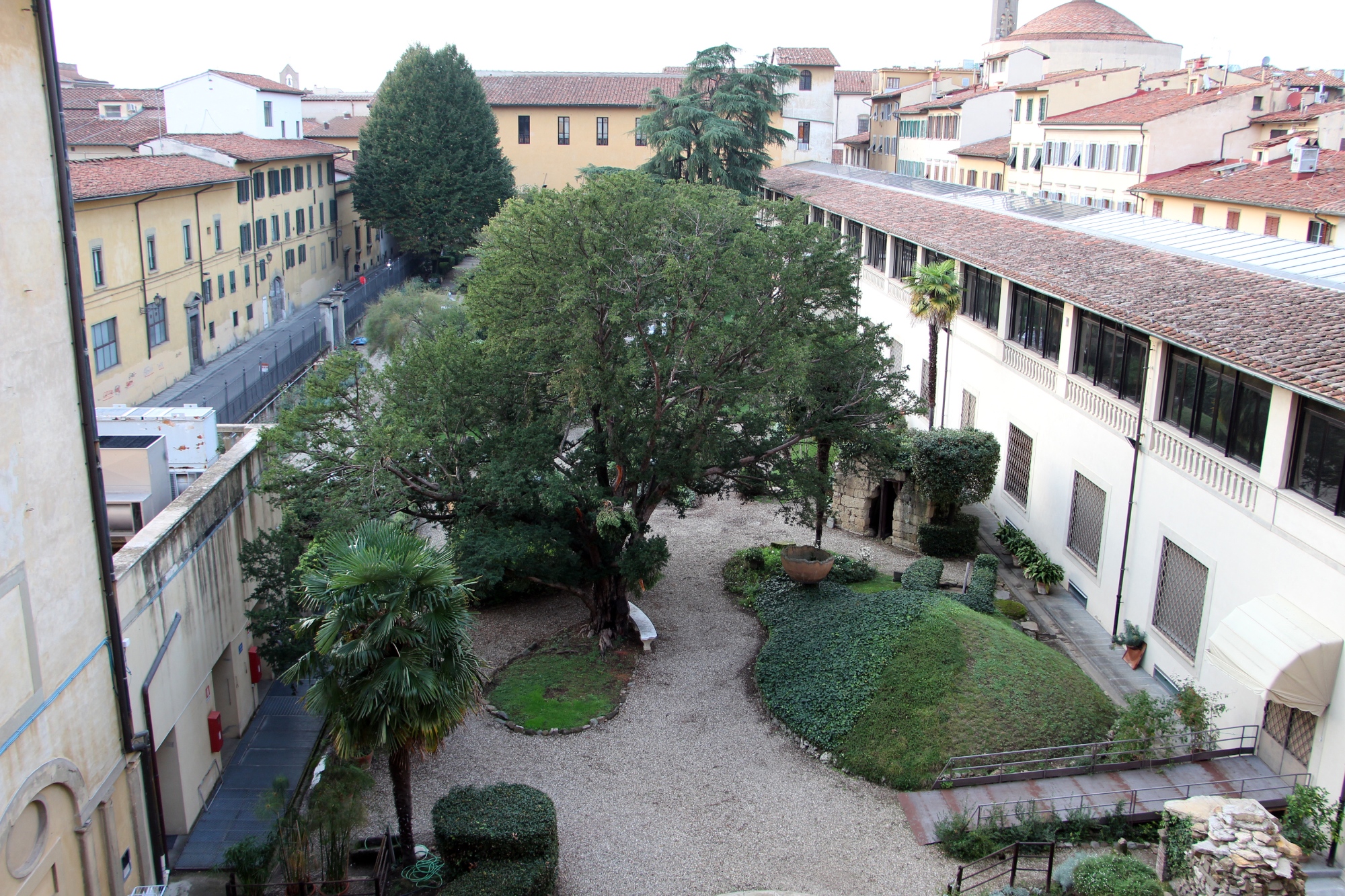 Jardin du musée archéologique national de Florence, avec des espaces verts entretenus et des allées constellées de statues et de pièces archéologiques en plein air, parmi les arbres et les plantes ornementales.