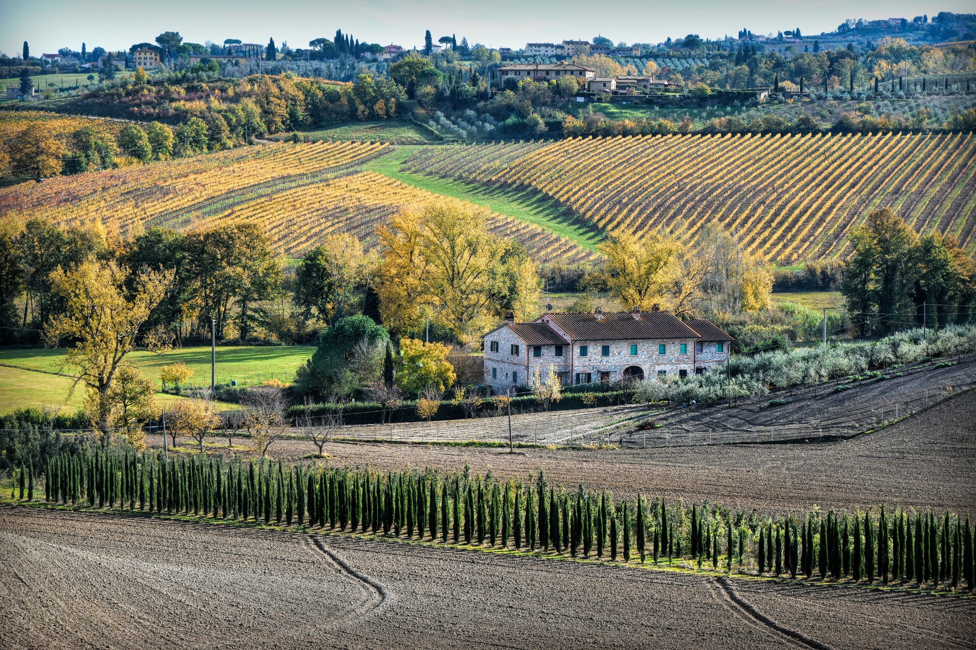 Paysage rural le long de la Via Francigena toscane : une ferme en pierre nichée au milieu des champs labourés, des rangées de cyprès et des vignobles dorés.